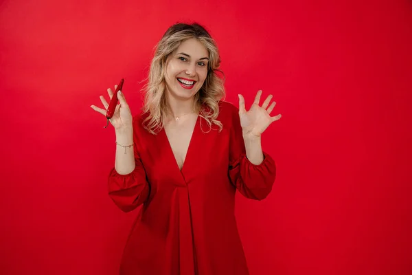 Laughing glad blond woman in red dress holding hot pepper in hand, shaking arms and gesturing, looking at camera on red isolated studio background. Hot news, sales, half price, banner ad. Copy space