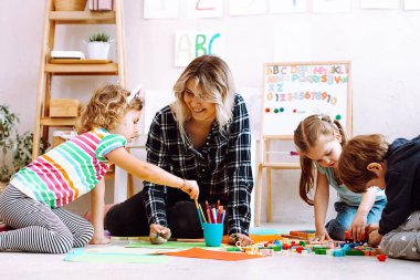 Portrait of young joyful woman teacher sitting near colored pencils, different toys with playing children, looking at serious little girl taking pencil in bright classroom. Education, forwardness.