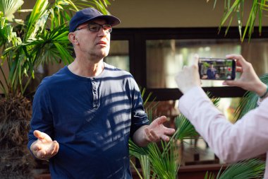 Portrait of serious middle-aged man blogger traveller influencer standing at palm trees in cafe, spreading hands, sharing experience, recording educational video blog with smartphone held by woman.