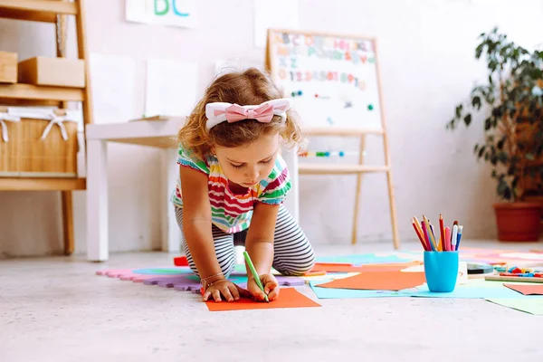 Portrait of pretty little girl with short fair hair wearing T-shirt, trousers sitting near magnetic board with numbers, alphabet, drawing with pencil on red sheet of paper around colorful papers. Art.