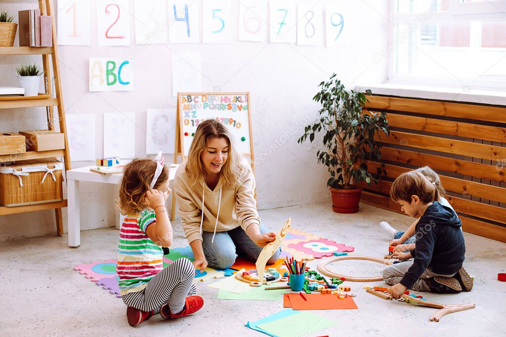 Portrait of young cheerful woman teacher sitting near colorful pencils, different toys with concentrated children, showing picture to little girl in bright classroom. Education, forwardness, crafts.