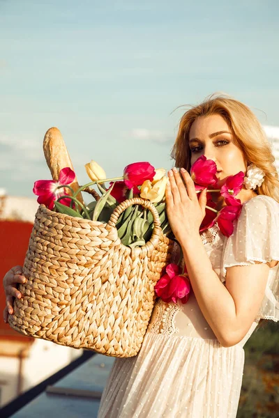 Attractive girl in romantic dress holding big wicker basket of fresh pink tulips and french baguette closeup, sky background. Paris style, relaxation on roof, cityscape, sky, floral composition.