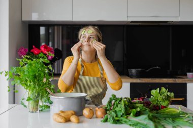 Funny and calm blonde woman with cucumber slices on face, cooking and eating in the kitchen. Female cooking natural and ecological organic eco food on weekend. Treatment and cosmetology at home. 