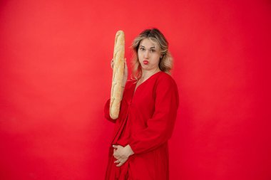 Playful funny blonde woman in red dress holding long french bread baguette and touching swollen belly on red studio isolated background. Dieting, weight loss and burning of calories and fat in summer