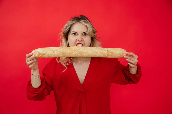 Hungry aggressive blonde woman in red dress holding biting long french bread baguette in hands on red studio isolated background. Advertisement of food, snacks, tasty, delicious food. Copy space
