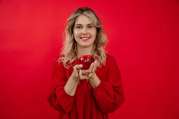 Young cheerful, merry, blissful blonde woman holding and presenting to camera fresh organic apple in two hands with smile on red isolated background. Healthy lifestyle, healthy eating. Copy space