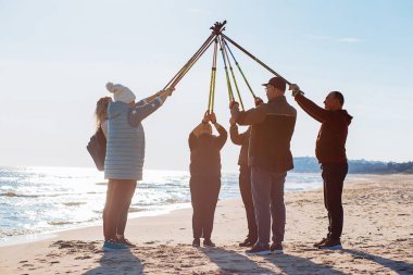 Group of people different ages in warm clothes in camp making Nordic walking crossing trekking sticks together in air against sea waves at sandy beach. Exercises activity for senior and mature people