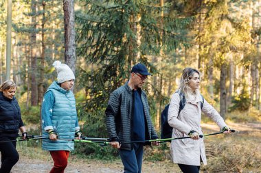 Side view of female coach, campers make Scandinavian walking one by one, hold trekking poles in the forest. Steps and breathing technic good posture. Exercise education. Healthy and active weekend