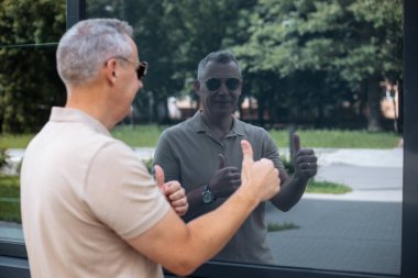 Portrait back view of senior smiling man in sunglasses walking on street along modern office building and looking on silhouette reflection in window. Show finger up, enjoy, like, approve.