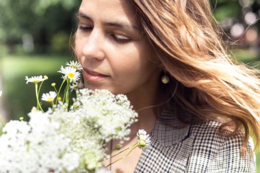Portrait closeup of young charming woman smelling and enjoying aroma of bouquet plucked white wild flowers. Walking in park on sunny day, outdoor relaxation, beauty of nature. Allergy free people.