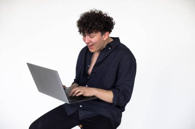 Young curly guy in total black look sitting on chair and playing computer games on modern laptop, gray studio background. Absorption into game, gambling addiction. Modern Internet reality.