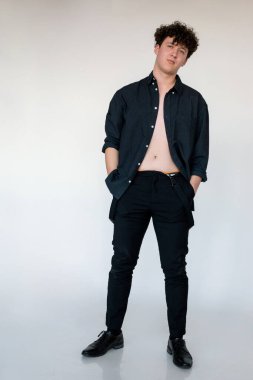 Full length photo of hot young curly guy in black unbuttoned shirt with naked torso, gray studio background. Trendy casual clothes for men, basic shirt. Comfortable fashion wear for every day.