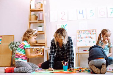 Kids and educator folding lego and drawing with colored pencils and markers sitting on floor in playroom from below view. Interesting lesson for kindergartners developing creation and intelligence.