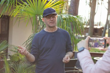 Portrait of serious middle-aged man blogger traveller vlogger standing at palm trees in cafe, making content for blog, sharing experience, recording educational video with smartphone held by woman.