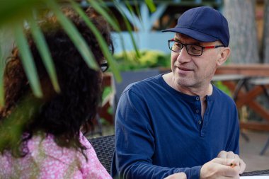 Portrait of handsome middle-aged man wearing blue cap, sweatshirt, glasses, sitting at table in cafe park outdoors, holding pen ready to write note, discussing with woman. Education, communication.