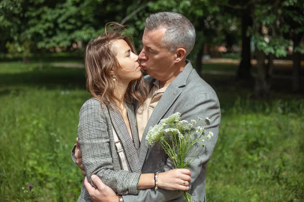 Portrait of cute couple hugging and kissing in green park on sunny day, nature background. Adult man and young woman walking with bouquet of wild flowers outdoors. Romantic date, true love, feelings.