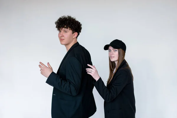Young couple standing sideways with arms bent at elbows in black blazers, gray studio background, copy space. Curly smiling guy posing with attractive girl in casual cap. Unisex clothes, basic wear.