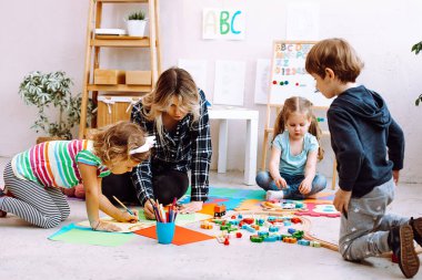 Young concentrated woman teacher sitting on floor near sheets of paper, colored pencils, different toys with pupils children, showing explaining how to draw with pencil in bright classroom. Education.