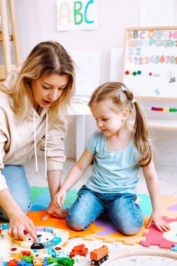 Portrait of beautiful little girl wear T-shirt, jeans sitting near colorful papers, different toys with young woman teacher teaching how to play with wooden parts of clock with numbers in classroom.