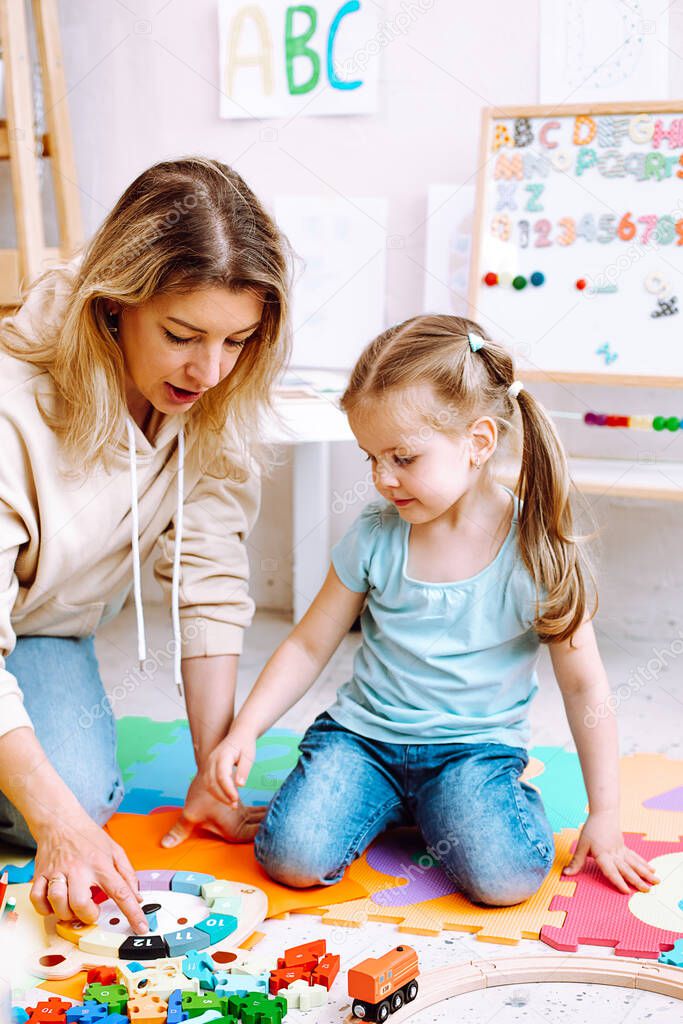 Portrait of beautiful little girl wear T-shirt, jeans sitting near colorful papers, different toys with young woman teacher teaching how to play with wooden parts of clock with numbers in classroom.