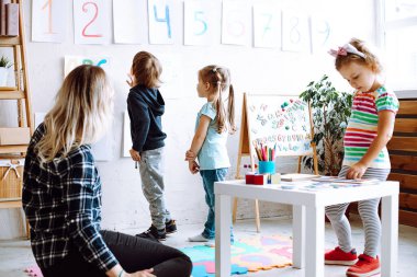 Group of adorable children pupils studying colorful numbers hung on wall with help of young teacher woman in bright classroom. Pretty little girl playing board games on table. Crafts, early education.
