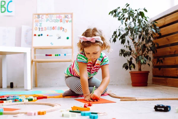 Portrait of cute little girl with short fair hair wearing T-shirt, trousers, sitting near magnetic board with numbers, alphabet, drawing outline of hand on red sheet of paper around different toys.