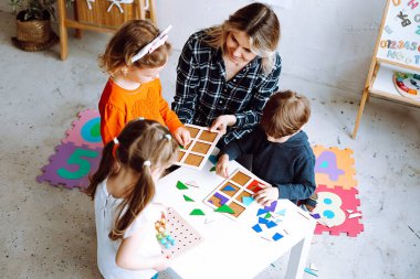 Top view of group of preschoolers and teacher. Children sitting around white table in bright classroom. Young blond woman helping pupils to play board games, collecting geometric figures. Forwardness.