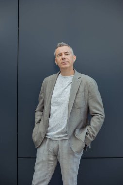 Portrait of confident aged gray haired businessman standing with hands in pants pockets and raised head in stylish suit on gray background. Entrepreneur, well dressed man, successful business.