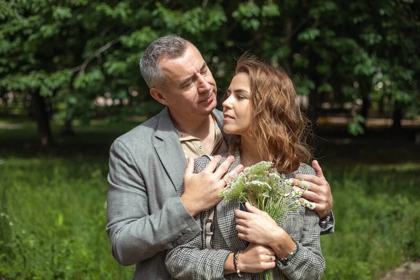 Portrait of loving couple gently hugging in green park on sunny day, nature background. Adult man and young woman walking with bouquet of wild flowers outdoors. Romantic date, true love, feelings.