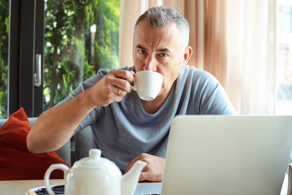 Aged handsome business man sitting in cozy cafe, drink tea and working on laptop. Implementation of modern technologies into urban lifestyle. Remote job, working time, freelance, busy people.