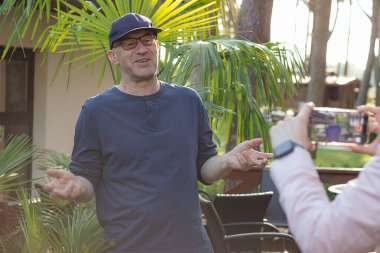 Portrait of smiling middle-aged man blogger influencer standing at palm trees in cafe, spreading hands, sharing professional skills, recording educational video blog with smartphone held by woman.