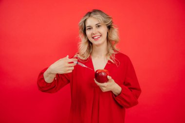 Smiling attractive woman hand with syringe applying injection to fresh juicy red apple on red isolated background. Beauty cosmetology rejuvenation industry. Botox injection treatment. Copy space