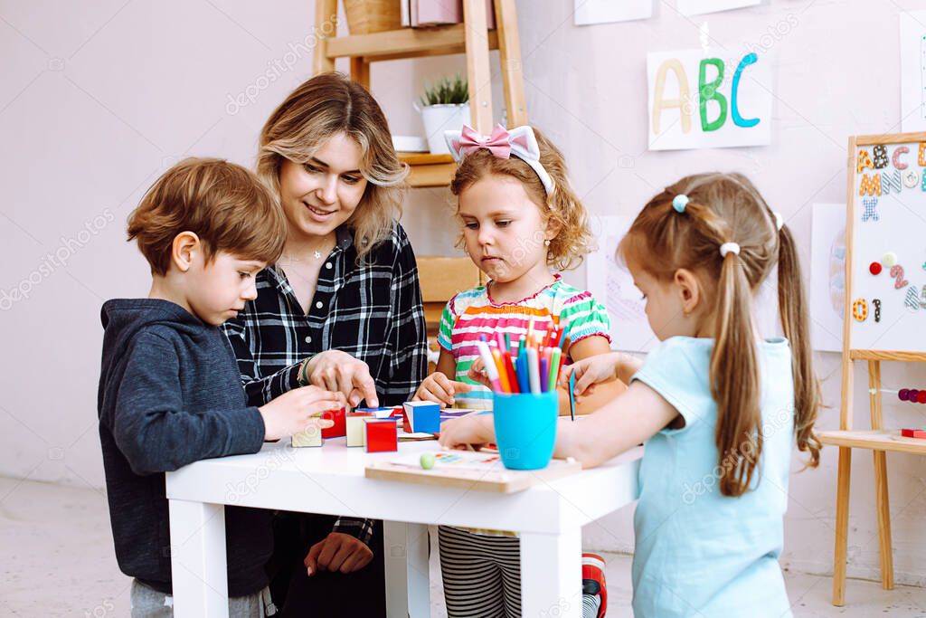 Portrait of young cheerful woman sitting at table with colored pencils, board games around children playing. Teacher explaining pupil boy how to complete task in bright classroom. Early development.