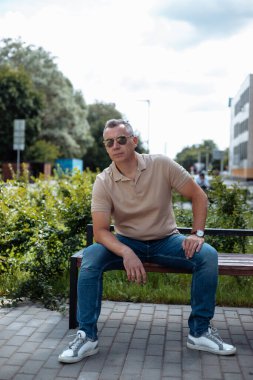Portrait of senior brutal man in sunglasses sitting on bench in landscaped house yard and resting on sunny day among green plants. Urban lifestyle, free time, outdoor recreation, summer leisure.