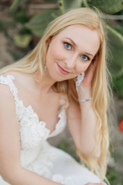 Portrait of young happy gorgeous blue-eyed woman bride titivating long fair hair wearing white wedding embroidered dress, sitting near green huge leaves of opuntia cactus. Wedding. Vertical, close-up.