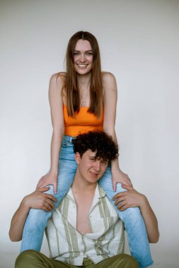 Rejoicing young couple in bright summer clothes, gray studio background, copy space. Cute girl in jeans sitting on shoulders of happy guy in unbuttoned shirt. Casual women and men wear for vacation.