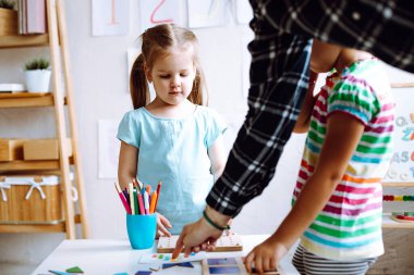 Two attentive cute little girls pupils standing at white table with colored pencils, playing board games in bright classroom. Teacher helping showing how to play children. Crafts, early development.