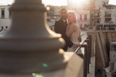 Side view of attractive wedding couple standing at steel railing of bridge at sunset. Young woman bride with long wavy fair hair wearing white wedding dress, embracing with groom in suit. Wedding. 