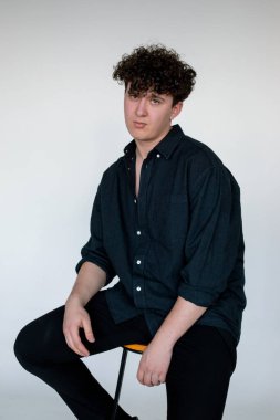 Beautiful young curly guy in total black look sitting on chair, gray studio background. Trendy casual clothes for men, basic shirt. Comfortable fashion wear for every day, street style clothing.
