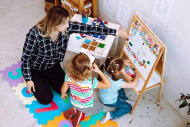Top view of young woman teacher sitting on floor near table with stationary, toys with girls pupils children, showing explaining alphabet numbers in classroom. Early education, forwardness, preschool.