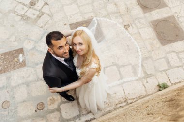 Top view of joyful wedding couple standing on pavement on street, looking at camera. Young woman bride with long fair hair wearing long white wedding dress, bridal veil, embracing with groom in suit.