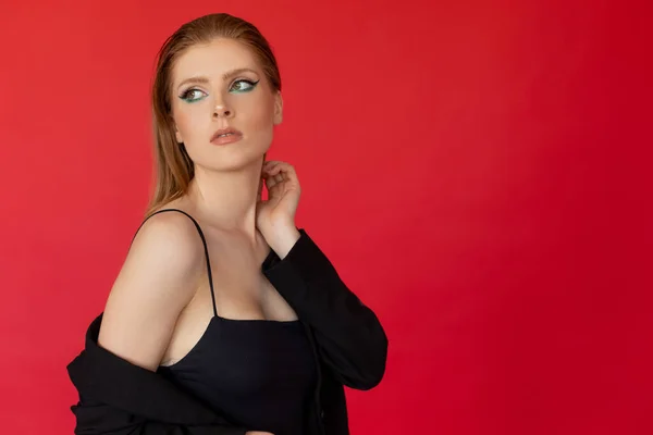 Portrait of young well-groomed woman with long ginger hair, bright make-up wearing black dress with straps, cardigan with bare shoulder, touching neck with hand on red background, looking aside.