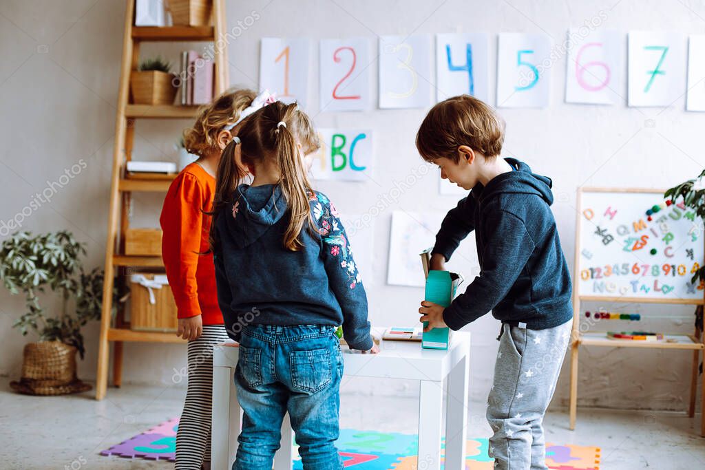 Group of wonderful children preschoolers studying playing in bright classroom. Pupils standing around white table near magnetic board with alphabet, numbers. Early development, preschool education.