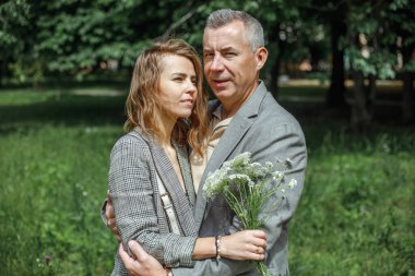 Portrait of beautiful couple gently hugging in green park on sunny day. Adult man and young woman walking with bouquet of wild flowers outdoors. Romantic date, true love, feelings, modern relations.