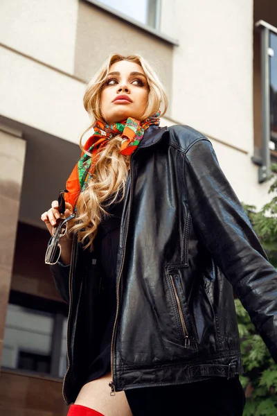 Blonde parisian girl with perfect makeup in stylish colorful headscarf, vintage leather jacket holding in hand sunglasses walking down street. Paris style, cityscape, urban lifestyle, fashion look.