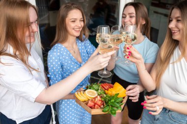 Portrait of young joyful amazing women holding vegetables, yellow peppers, green leaves of basil, oranges on wooden tray, clinking glasses of white wine, standing near house. Friendship, party.