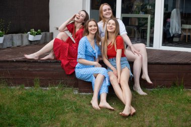 Group of young smiling gorgeous barefoot women sitting on wooden veranda near green grass, looking at camera, laughing, having fun, enjoying weather. Friendship, relationship, hen-party, summer.