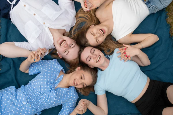 Top view of young laughing stunning women holding joining hands together, lying in circle on blue blanket with closed eyes, resting. Symbol of unity, support, collaboration, mutual help, relaxation.