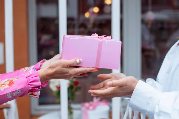 Cropped close up woman hands with manicure giving present pink box with ribbon as wrapped gift to other woman in white shirt, hand in hand. Celebration of holidays, birthday party against glass doors