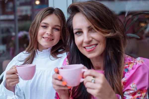 Calm smiling mother and daughter drinking coffee in outdoor cafe, spending time together on weekends. Pretty fashionable long haired woman and teenager on brunch with beverage. Mother day together